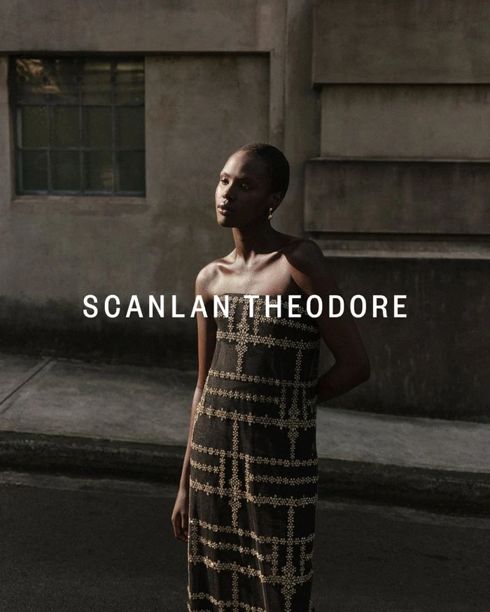 Model Achok Akoi wearing a patterned strapless dress standing outdoors against a textured architectural building exterior.
