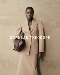 Model in camel-colored blazer and matching skirt holding a dark brown handbag against a textured concrete wall.