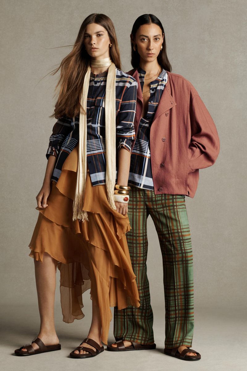 Two fashion models showcasing apparel in a studio setting. One wears a plaid shirt and layered skirt, the other wears a jacket and plaid pants.