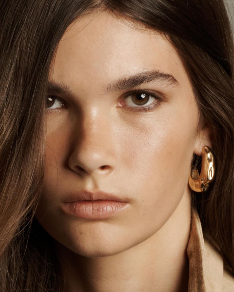 Close-up portrait of a young adult female with brown hair and eyes wearing a golden hoop earring and tan colored clothing.
