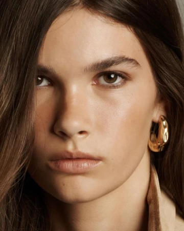 Close-up portrait of a young adult female with brown hair and eyes wearing a golden hoop earring and tan colored clothing.