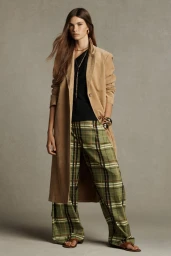 Model River Klein posing in a tan trench coat, black top, and green plaid wide-leg trousers against a grey seamless backdrop.
