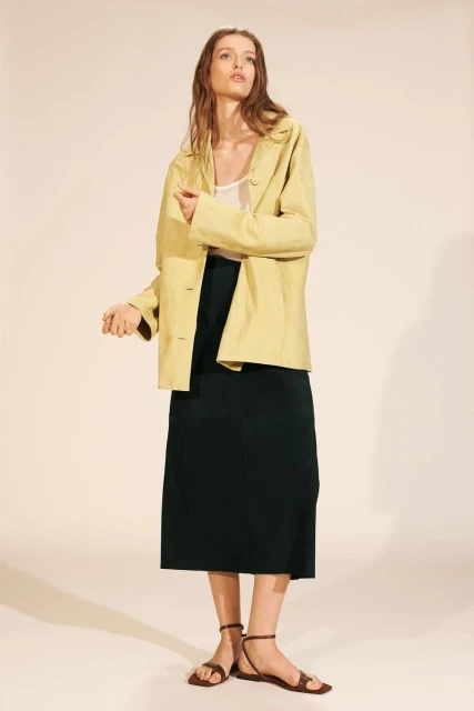 A young adult woman in a fashion pose wearing a pale yellow blazer, dark green skirt, and brown sandals on a beige background.