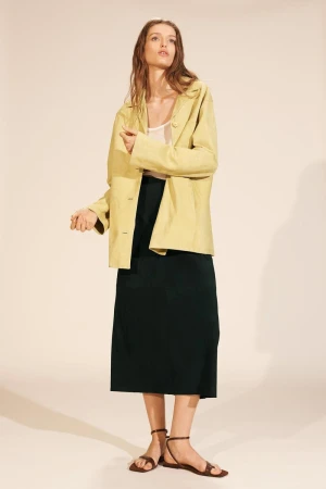 A young adult woman in a fashion pose wearing a pale yellow blazer, dark green skirt, and brown sandals on a beige background.