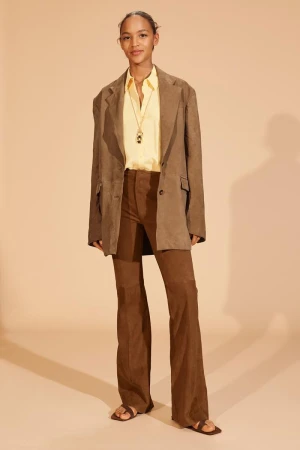 Fashionable woman poses in brown suede blazer, yellow blouse, brown suede pants, and sandals, standing against a light beige backdrop.