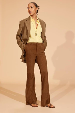 A woman models a yellow blouse and brown suede pants with matching blazer in a studio setting with a neutral tan background.