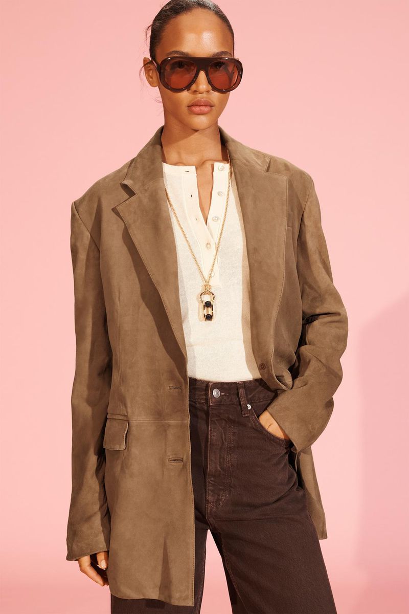 Stylish woman in a brown suede blazer and matching sunglasses posing against a pink background, showcasing a fashion-forward look.