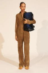 Model in brown suede jumpsuit holding a blue woven bag against a seamless studio background