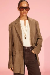 Model wearing oversized brown suede blazer, dark sunglasses, and pendant necklace against a pink backdrop