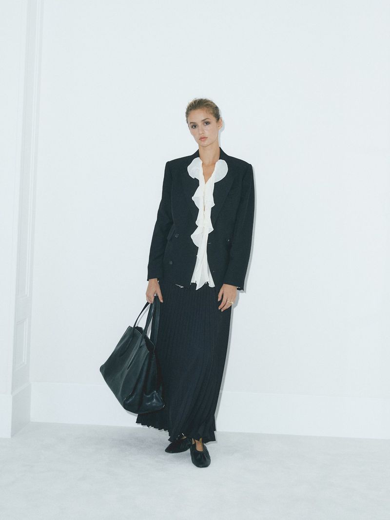 Fashionable woman in a black blazer, white ruffled blouse, and long skirt, holding a large black tote bag in a light studio.