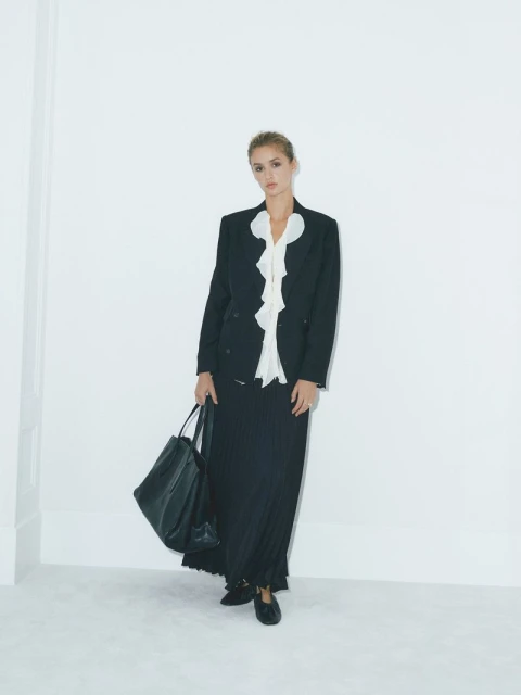 Fashionable woman in a black blazer, white ruffled blouse, and long skirt, holding a large black tote bag in a light studio.