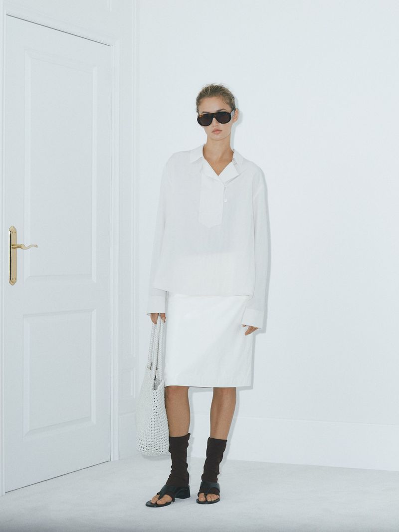 Stylish woman poses in a white shirt and skirt, accessorized with sunglasses and a tote bag, standing near a white door and wall.