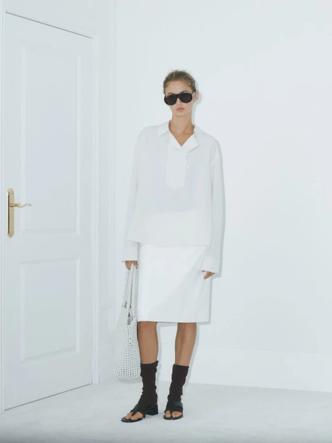 Stylish woman poses in a white shirt and skirt, accessorized with sunglasses and a tote bag, standing near a white door and wall.