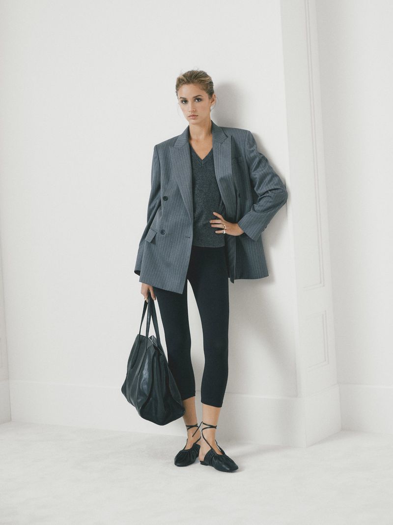 A woman poses in a gray blazer, black leggings, and flats, holding a large tote bag against a white wall for a fashion shot.