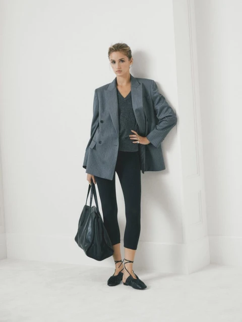 A woman poses in a gray blazer, black leggings, and flats, holding a large tote bag against a white wall for a fashion shot.