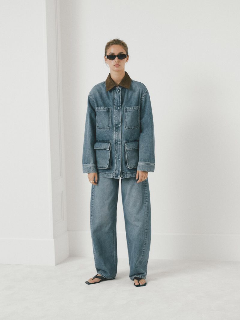 Woman in baggy denim jacket and jeans against a white wall. Fashion shot with neutral tones, complemented by sunglasses and dark sandals.