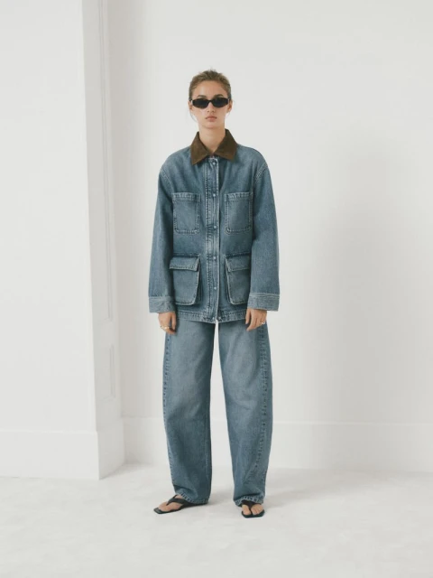 Woman in baggy denim jacket and jeans against a white wall. Fashion shot with neutral tones, complemented by sunglasses and dark sandals.