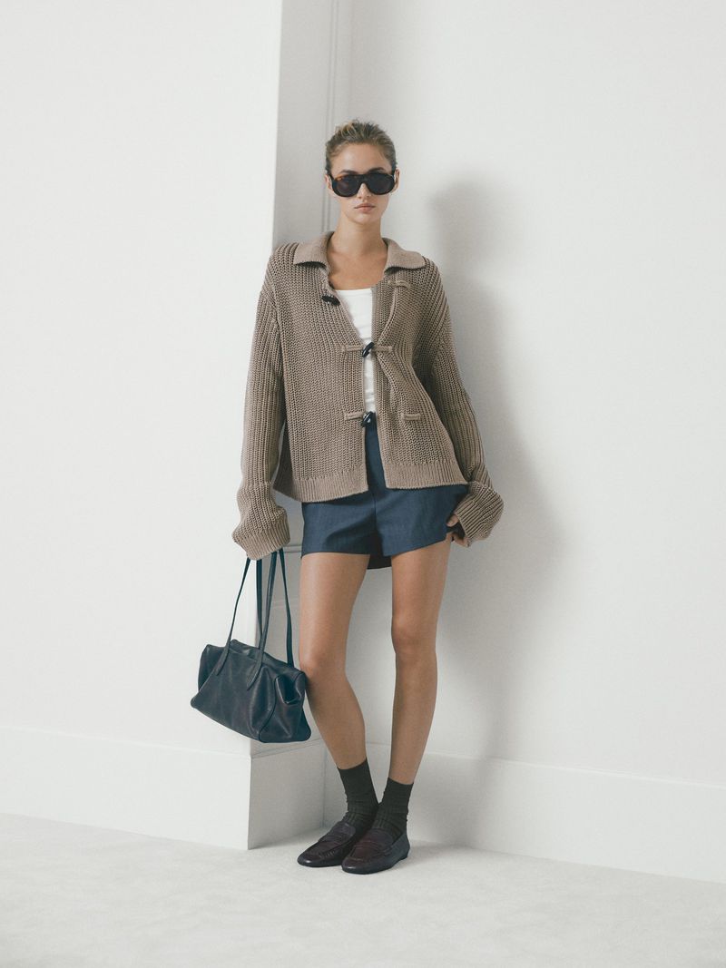 A slender woman poses against a white wall wearing a tan cardigan, blue shorts, dark socks and shoes, holding a black leather handbag.