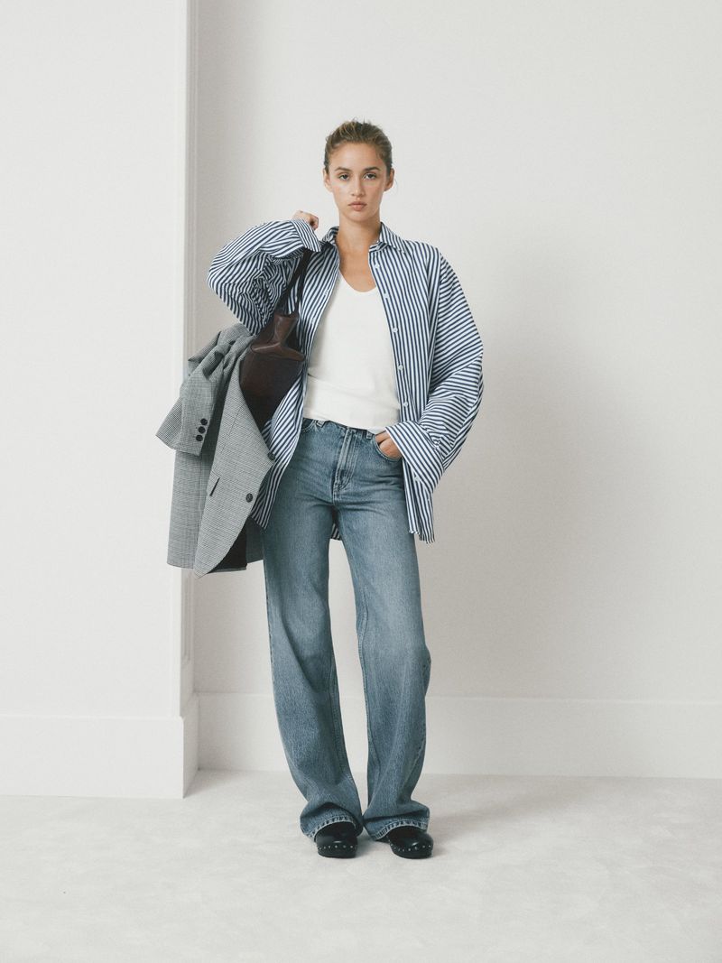 Woman in oversized blue striped shirt and wide-leg jeans poses in a bright studio, holding a grey blazer and brown shoulder bag.