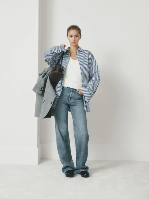 Woman in oversized blue striped shirt and wide-leg jeans poses in a bright studio, holding a grey blazer and brown shoulder bag.