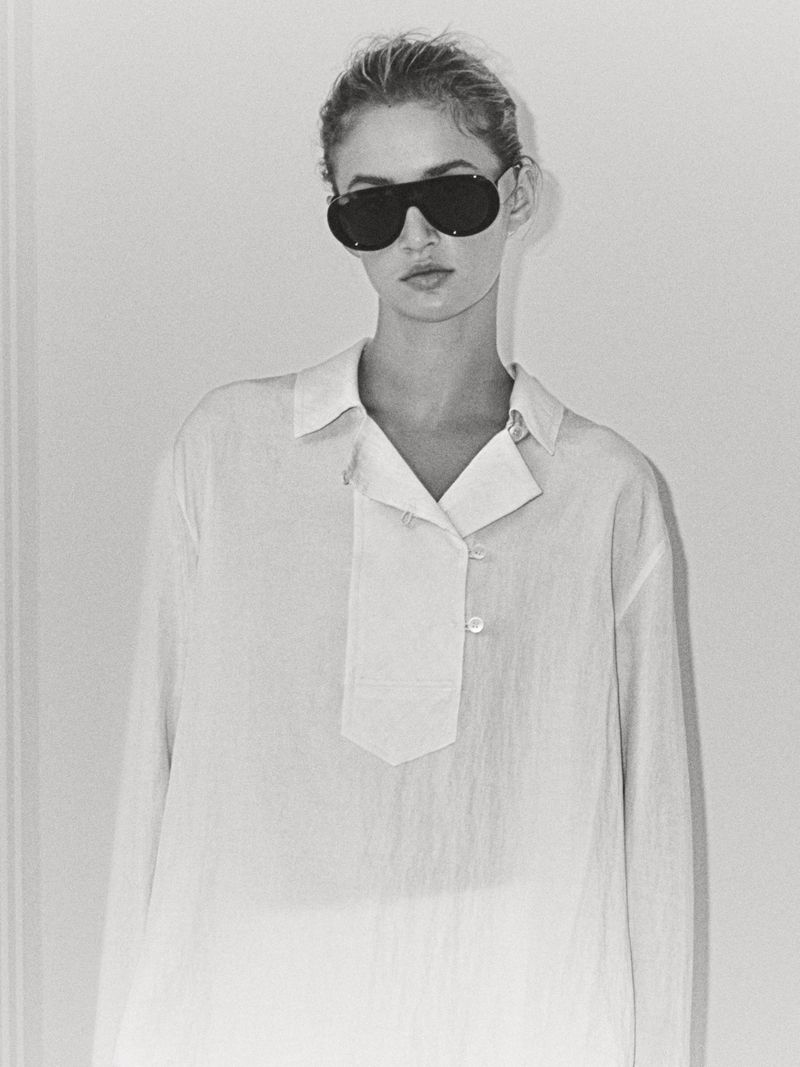 A young woman with sunglasses poses in a white button-down shirt against a white wall in a studio setting with diffused lighting.