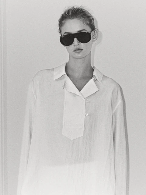 A young woman with sunglasses poses in a white button-down shirt against a white wall in a studio setting with diffused lighting.