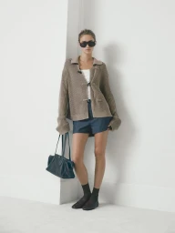 Model Quinn Mora wears a taupe knit cardigan, navy shorts, dark sunglasses, and black loafers against a white backdrop.