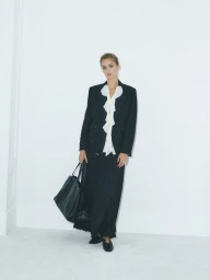 Model wearing a black blazer over a white ruffled blouse and black skirt, holding a large black handbag against a plain studio background.