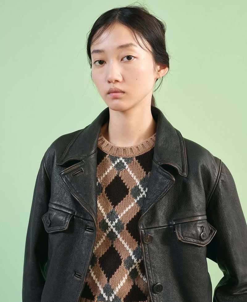 A young adult woman poses in a brown and black argyle sweater and black leather jacket against a light green backdrop in a medium shot.