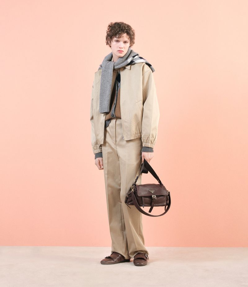 Male model wearing a khaki jacket and pants, grey scarf, and holding a brown leather bag, posing against a peach-colored background.