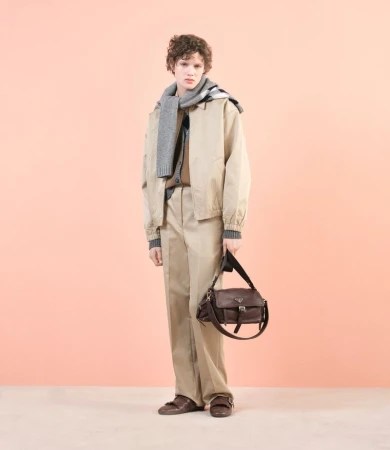 Male model wearing a khaki jacket and pants, grey scarf, and holding a brown leather bag, posing against a peach-colored background.