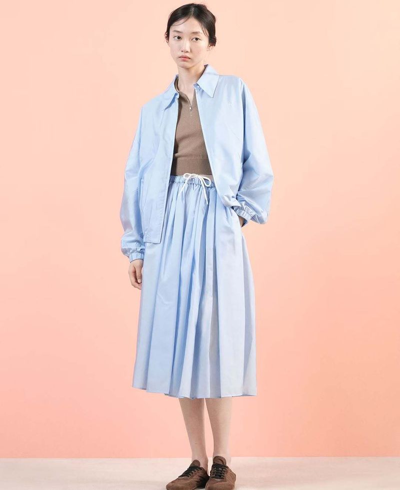 An Asian woman models a light blue jacket and skirt set with brown sneakers in a studio against a peach-colored background.