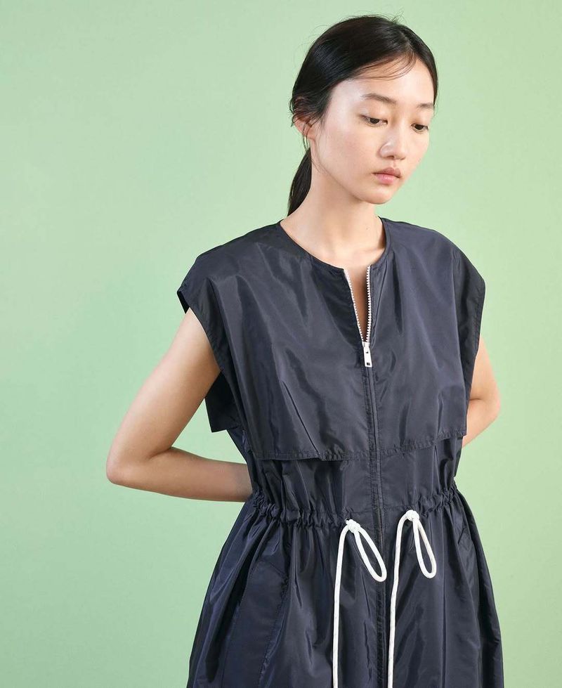Young Asian woman wearing a black nylon dress with a front zipper and white drawstring against a light green background in a studio.