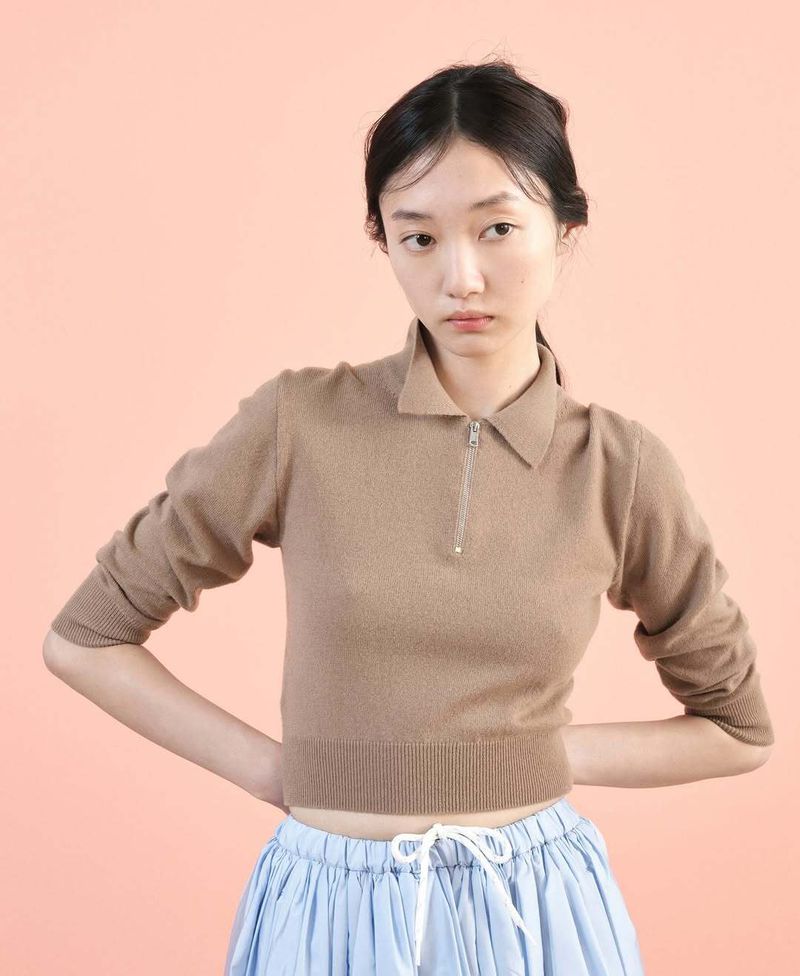 Young woman poses in a brown collared, zipper sweater and blue skirt against a salmon background in a studio portrait.
