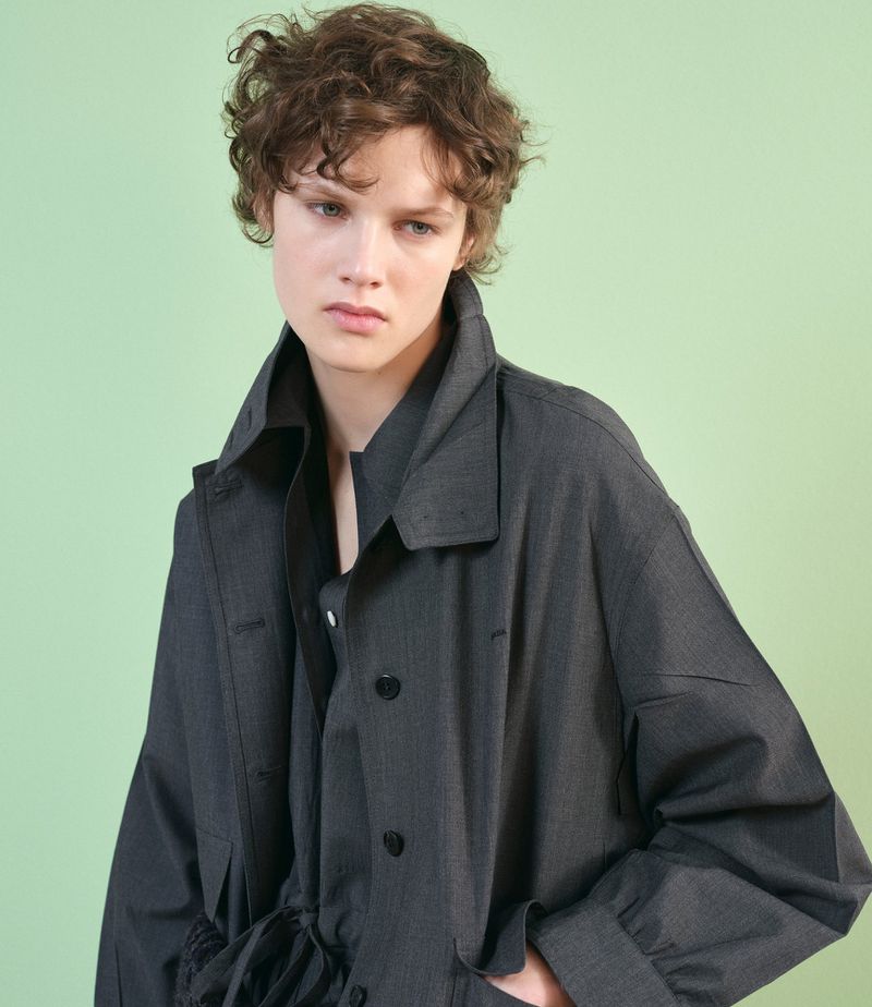 Young adult poses in a gray-blue button-down coat against a pale green backdrop, showcasing fashion and style in a studio setting.