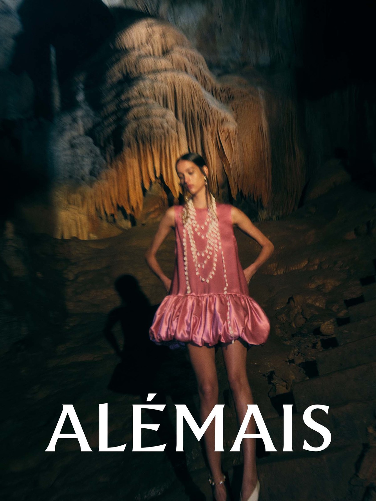 Fashion shot in a cave featuring a model in a pink dress and multiple strands of pearls, with dramatic lighting and a geological cave formation.