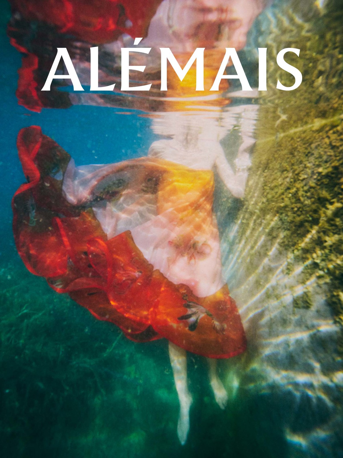 Underwater shot of a person in a flowing red and white dress next to a rock, with sunlight filtering through the water.