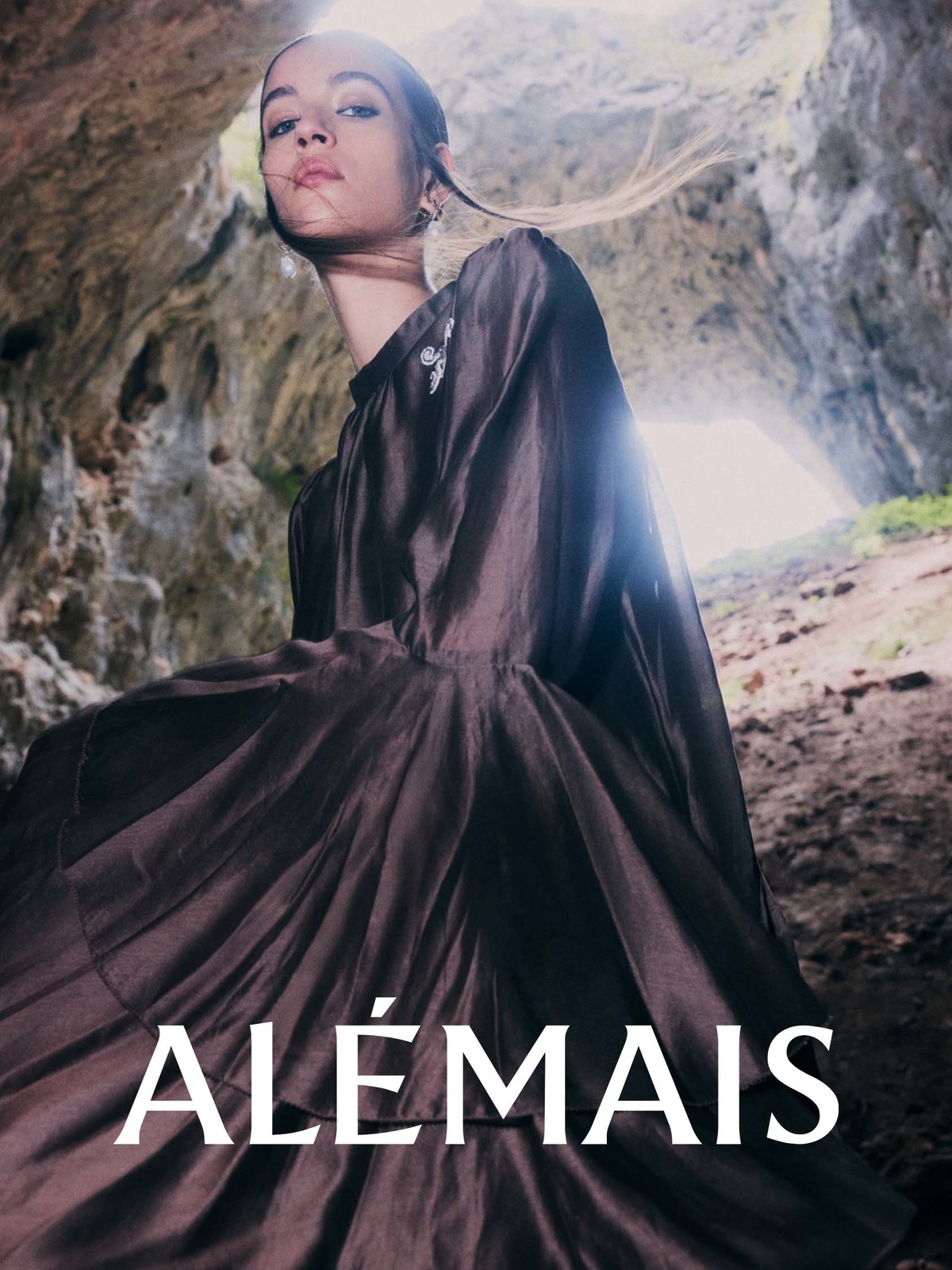 Woman in dramatic brown dress poses inside a stone cave, illuminated by bright light, with the brand name 'ALÉMAIS' visible.