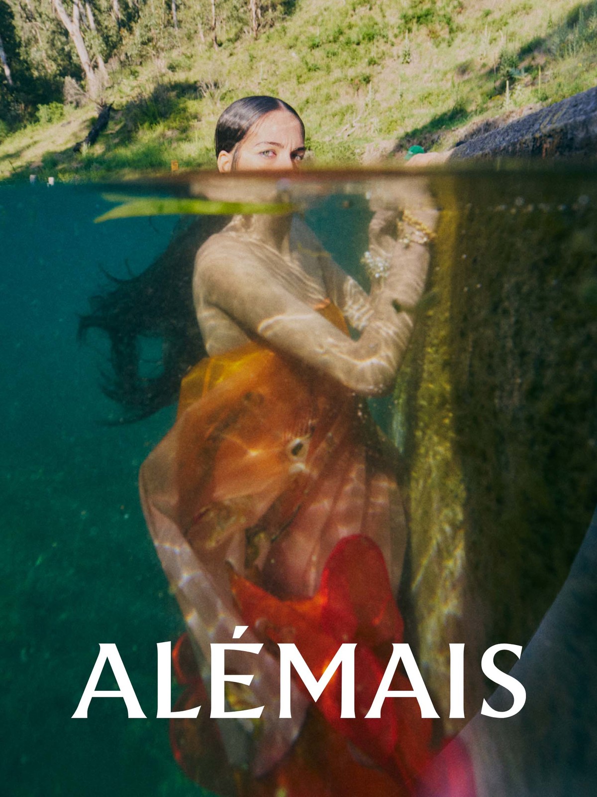 Woman underwater wearing orange and red dress. Half of image shows lush green trees and sky. Fashion photography, natural light.
