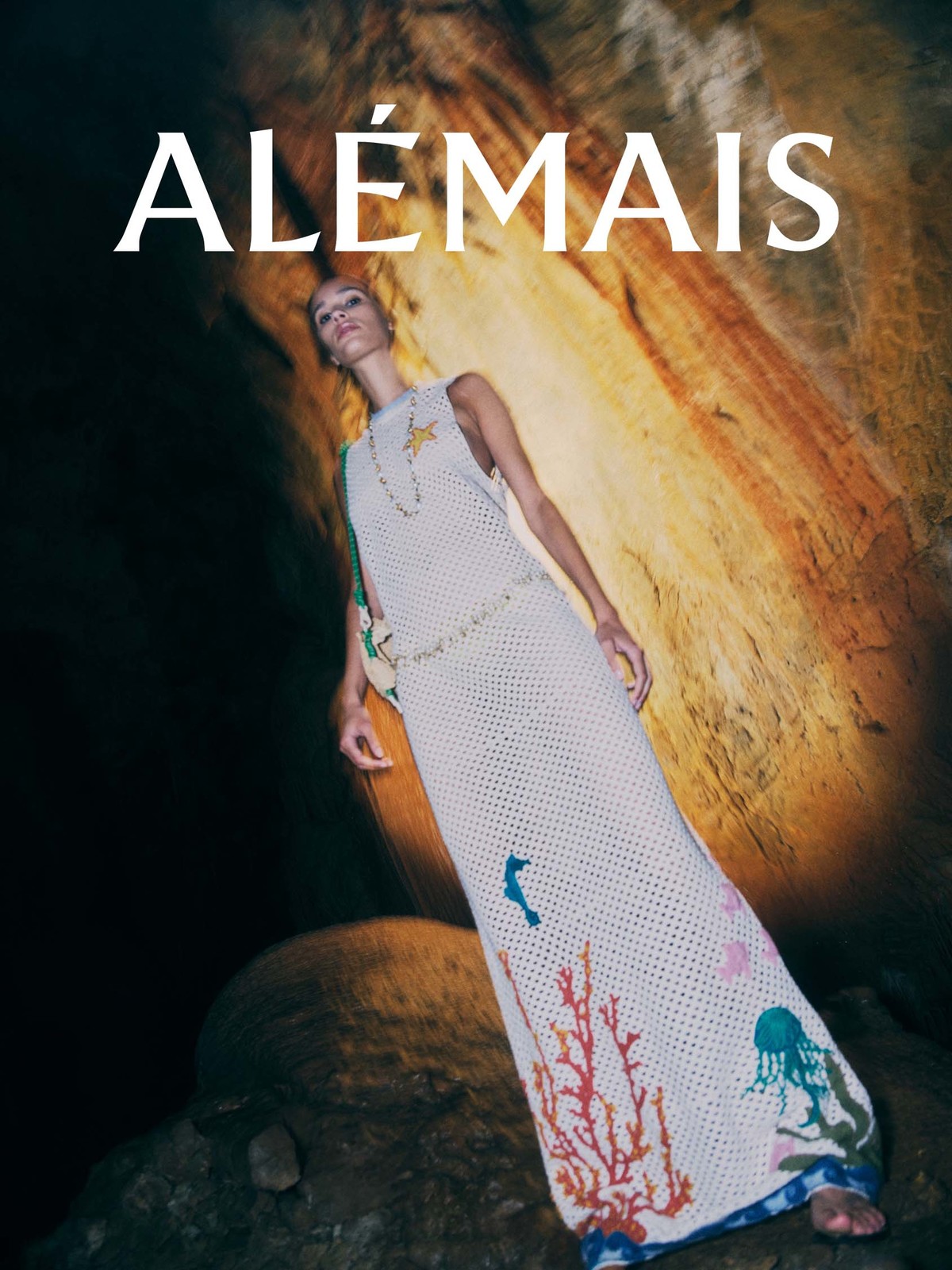 Model wearing a long white knit dress with marine life motifs inside a rocky cave