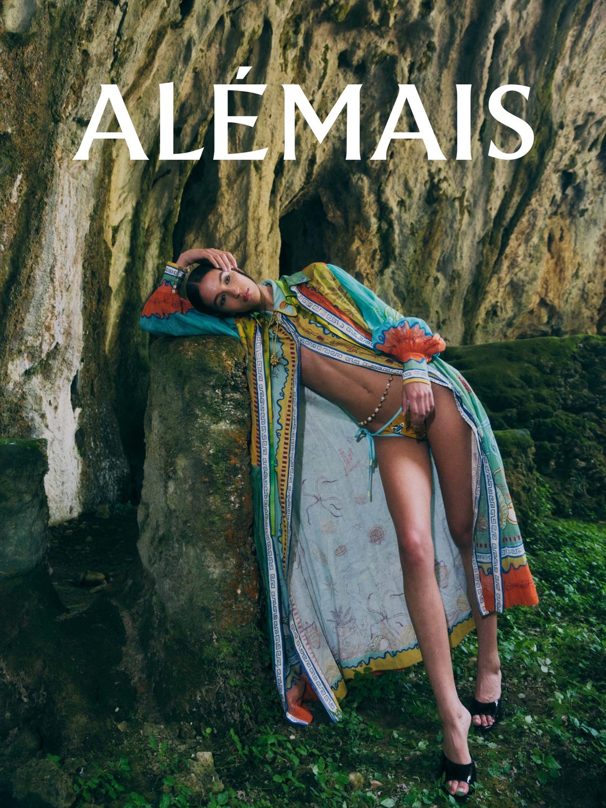 Woman poses wearing a patterned duster and bikini near a stone cave in a fashion shot. She wears black heels in the outdoor setting.