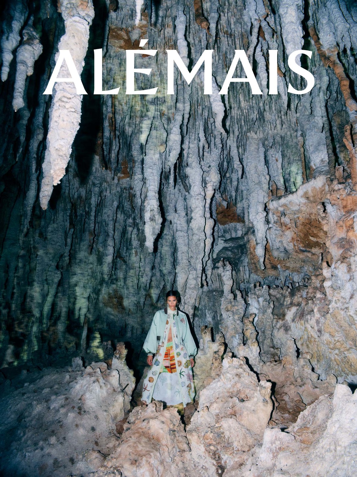 Woman in printed coat and matching pants poses in a large cave with stalactites, under the ALEMAIS logo in a fashion editorial.