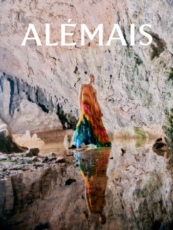 Model wearing a colourful patterned halterneck maxi dress standing inside a natural cave with a shallow pool reflecting her figure