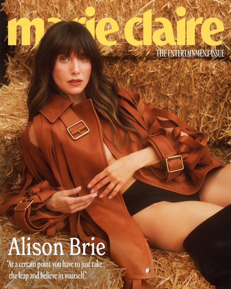 Magazine cover featuring Alison Brie, wearing a rust-colored jacket, and black bottoms, lying on hay in a portrait layout.