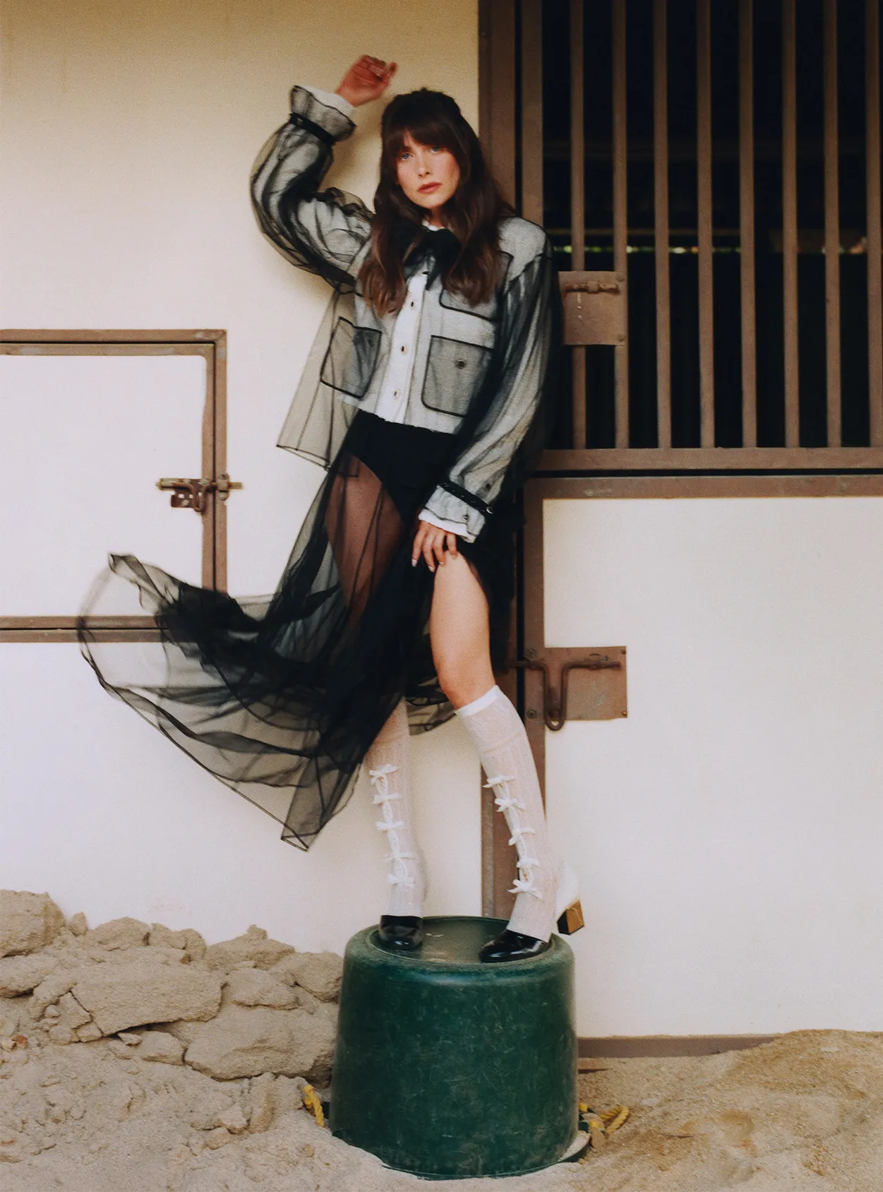 A woman poses on a bucket in a sheer black dress and jacket, accessorized with long socks with bows in a horse stable.