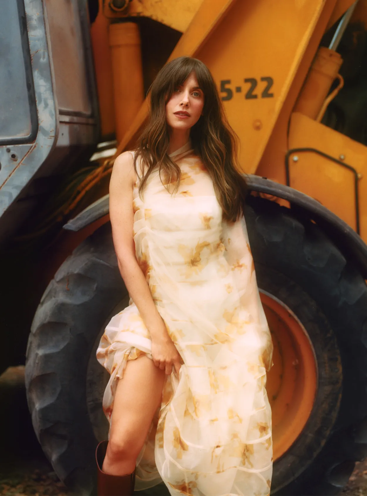 A woman in a flowing dress and boots stands in front of a yellow construction vehicle with large tires in an outdoor setting.