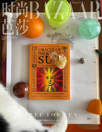 Overhead shot of 'Oracle of the Radiant Sun' book alongside objects including a crystal, orange, and a clear green cup on a white surface.