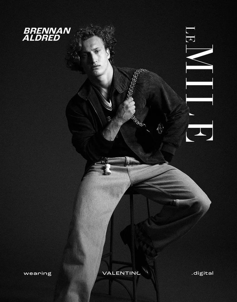 Monochrome portrait of a male model seated on a stool wearing a jacket, jeans, and a chain bag for a fashion editorial.