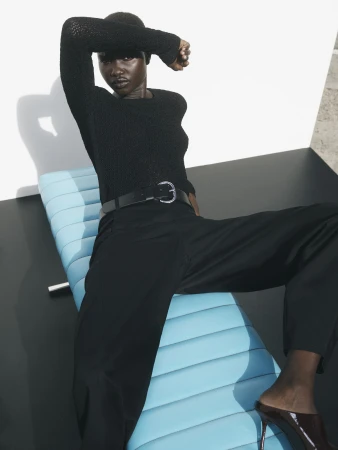 Elegant studio shot: African woman poses on sleek blue chaise lounge, wearing a black knit top and trousers with pointed-toe heels.