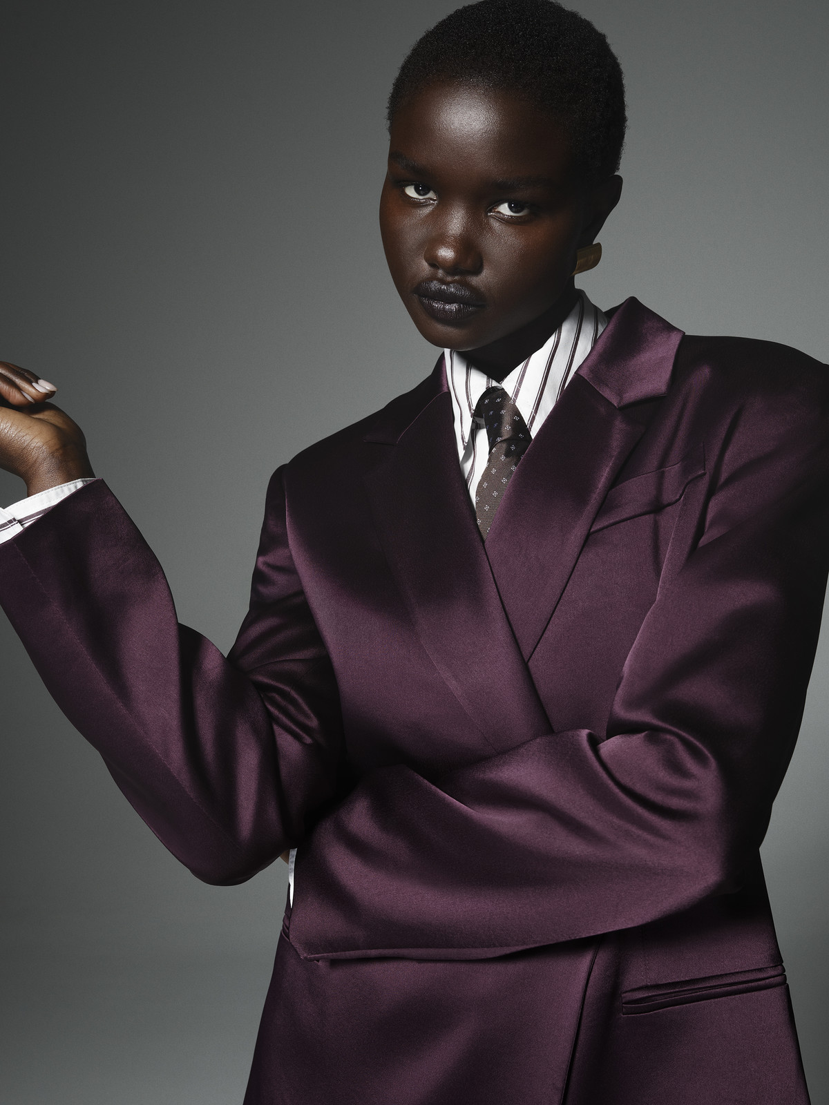 Dark-skinned model poses in a studio wearing a purple satin suit jacket, white striped collared shirt, and a dark tie, against gray backdrop.